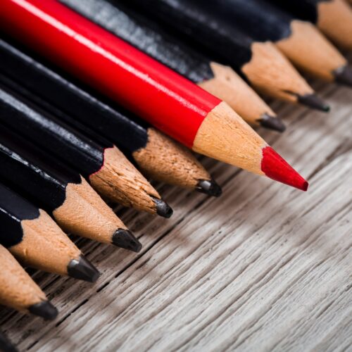 Red pencil stands out from the crowd of black pencil on a wooden white background. Leadership, uniqueness, independence, initiative, strategy, disagree, think, different, concept of business success